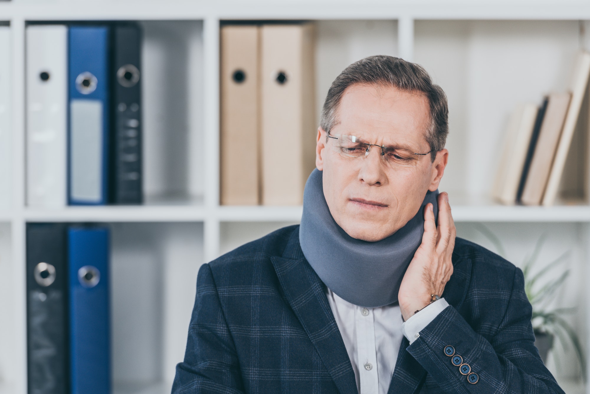 man in neck brace sitting, feeling pain and holding neck in office, compensation concept